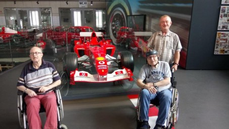 zwei Männer im Rollstuhl vor einem Formel 1 Rennwagen. Ein Mann steht hinter einem der Rollstühle.