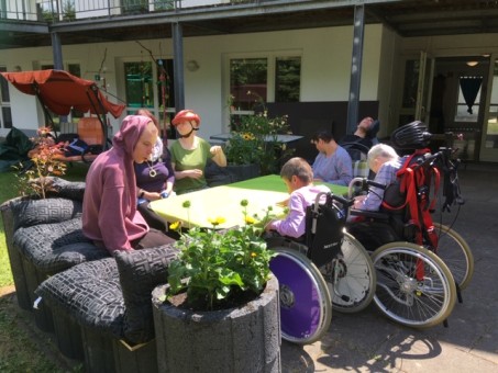 sechs Personen sitzen um einen Tisch im Garten. Einige Perssonen sitzen im Rollstuhl.