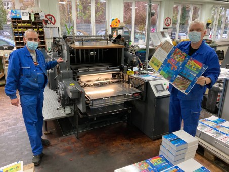 Zwei Männer in Blaumännern stehen rechts und links von einer Druckmaschine, der eine von Ihnen hält einen Druckbogen mit Bildern in den Händen